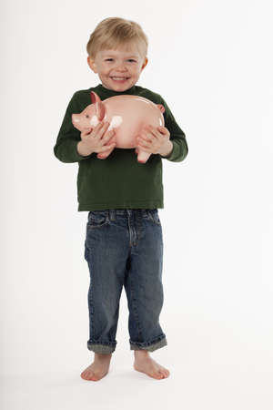 A young, barefoot boy is standing and smiling while holding a piggy bank. Vertical shot.の写真素材