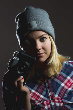 Portrait of hipster girl posing with retro camera wearing checkered shirt and winter hat on dark...