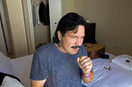 Hispanic man sitting on bed in his bedroom and holding his hand over his mouth to cough.の写真素材