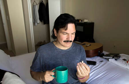 Hispanic man sitting on his bed looks at pill before taking it with a beverage.の写真素材
