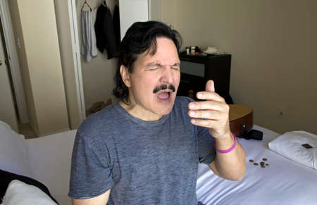 Hispanic man sneezes while sitting on bed in bedroom.