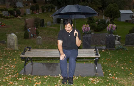 Hispanic man sitting on bench with lowered head and holding umbrella in cemetery.の写真素材