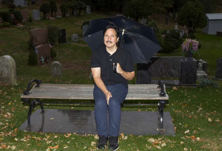 Hispanic man sitting on bench and holding umbrella in cemetery.の写真素材