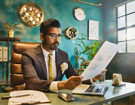 Young indian businessman in suit and glasses sitting at desk in officeの素材