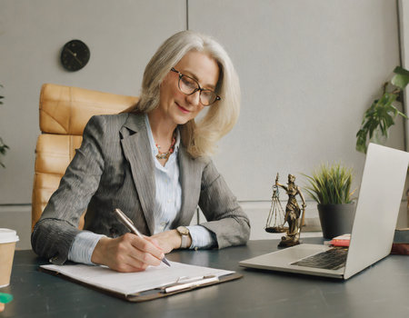 attractive mature businesswoman in eyeglasses writing in notebook at workplaceの素材