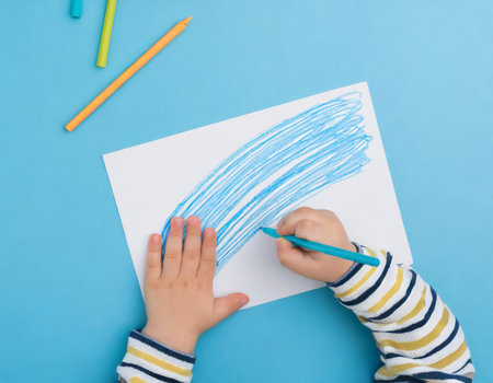 Top view of a child's hand drawing with colored pencils on a white sheet of paperの素材