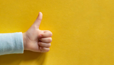 Child's hand shows thumbs up on a yellow background. Copy space.の素材