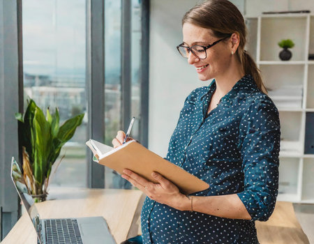 smiling pregnant businesswoman writing in notebook and looking away in officeの素材