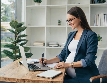 smiling pregnant businesswoman writing in notebook while sitting at table in officeの素材