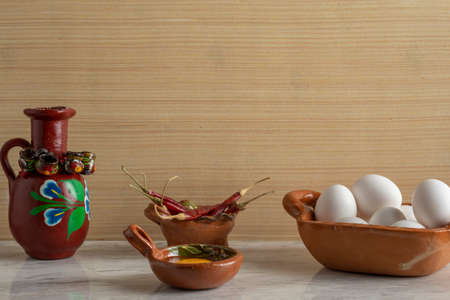 Egg in a clay bowl, arbol chili peppers and a decorated clay jug, on a marble bar and wooden...