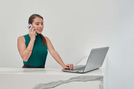 Receptionist making cell phone call while checking his laptop, in a cubicle with marble finish and white walls.の写真素材