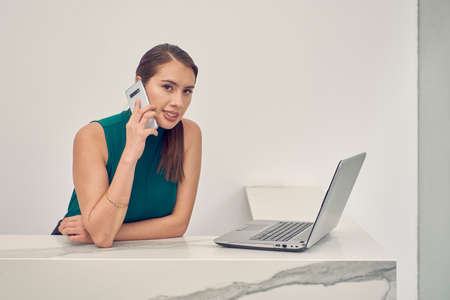Receptionist making cell phone call while checking his laptop, in a cubicle with marble finish and white walls.の写真素材