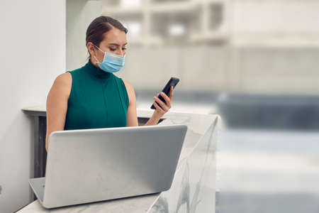 Executive woman checking and answering messages on her cell phone, wearing a mask due to normality...