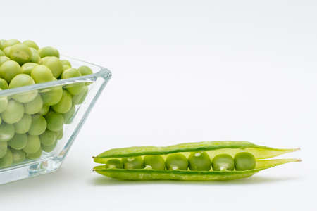Peeled peas in a glass container with some peas in the shell on one side on a white backgroundの写真素材