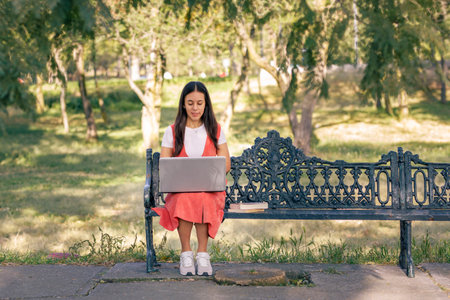Girl working in the park with her laptop, sitting on a bench with trees by the sides.の写真素材