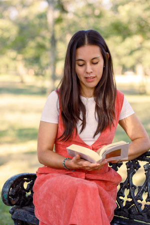 Young Latin woman, reading a book sitting on a bench in a park, studying outdoors.