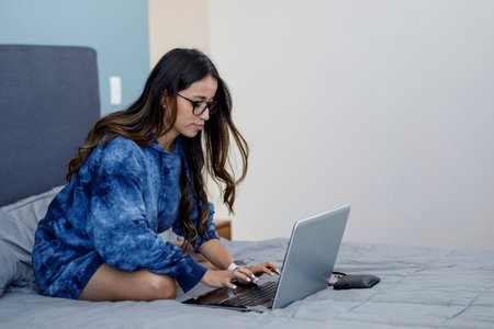 Young Latin woman working in her bedroom with a laptop wearing comfortable clothes. Home office concept for pandemicの写真素材