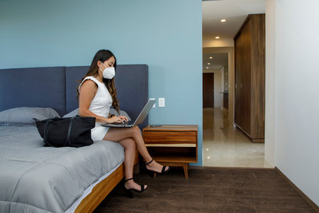 Young Latina businesswoman wearing a mask while working on her laptop, in her apartment bedroom doing home office.の写真素材