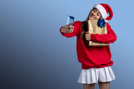 young latin woman hugging a gift with christmas clothes taking a selfie with her phoneの写真素材