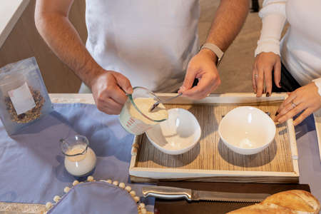 Hands emptying yogurt into a bowl with a spoon to prepare breakfast in a kitchen