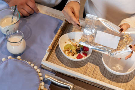 Womans hands emptying granola into a bowl with yogurt while preparing breakfast with her husband