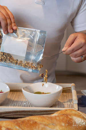 Mans hands emptying granola into a white bowl in a kitchenの写真素材