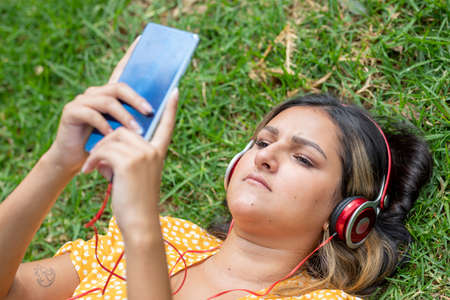 Woman lying on the grass listening to music while chatting on her mobile phoneの写真素材