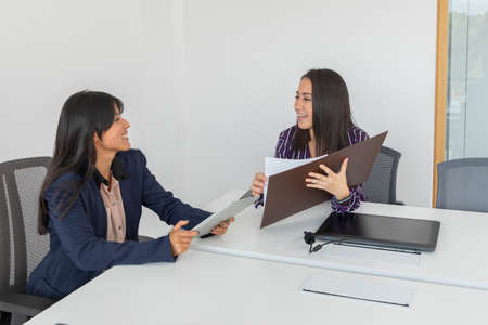 Two young Latina businesswomen review work documents in an office. Executive women signing important...