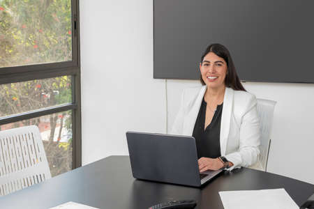 Portrait of a young business woman looking at the camera, in her office while working on her laptop.