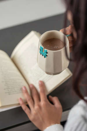 vertical shot of a Latin woman holding a cup of coffee, while reading a book in the morning