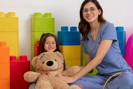 Pediatrician comforting smiling girl hugging teddy bear in colorful playroomの写真素材