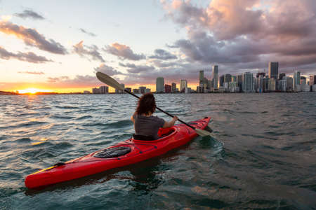 Adventurous girl kayaking in front of a modern Downtown Cityscape during a dramatic sunset. Taken in Miami, Florida, United States of America.の写真素材