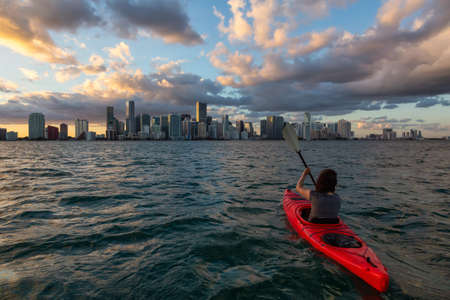 Adventurous girl kayaking in front of a modern Downtown Cityscape during a dramatic sunset. Taken in Miami, Florida, United States of America.の写真素材