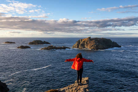 Woman in red jacket is standing at the edge of a cliff with open arms and enjoying the beautiful...