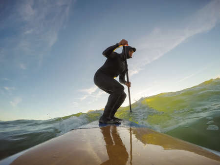 Adventurous Man Surfer on a paddle board is surfing in the ocean during a sunny sunset in Fall Season. Taken in Long Beach, Tofino, Vancouver Island, BC, Canada.の写真素材