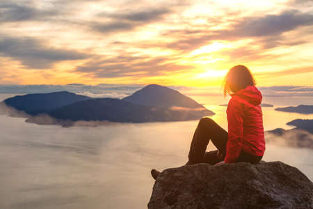 Adventurous Caucasian Girl sitting on top of a mountain during a colorful winter sunset. Taken on Tunnel Bluffs Hike, North of Vancouver, BC, Canada.の写真素材