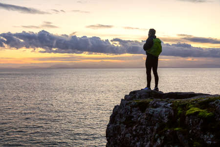 Adventurous Latino Male is standing on a rocky cliff by the Pacific Ocean during colorful cloudy sunset. Taken in Lighthouse Park, West Vancouver, BC, Canada.の写真素材