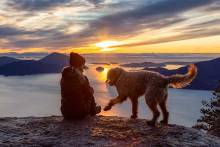 Adventurous Girl Hiking on top of a Mountain with a dog during a colorful sunset. Taken on Tunnel Bluffs Hike, near Vancouver and Squamish, British Columbia, Canada.