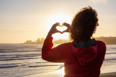 Long Beach, Near Tofino and Ucluelet in Vancouver Island, BC, Canada. Adventurous Girl making Hearth Shape with Hands with golden sunset on the Pacific Ocean Coast in the background.の写真素材
