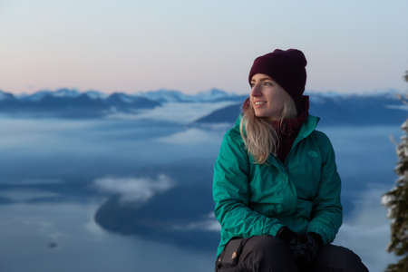 Vancouver, BC, Canada - November 25, 2019: Adventurous Blond Caucasian Girl sitting on top of a snow covered mountain during a colorful winter sunset.のeditorial素材