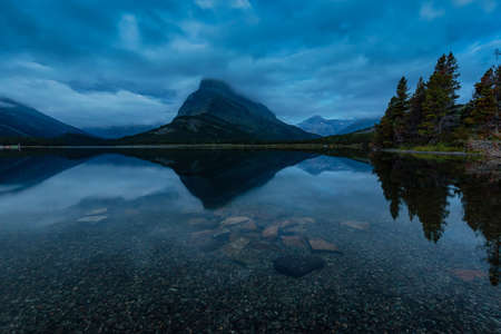 Beautiful View of the reflection in the water during a cloudy morning sunrise in the American Rockies. Taken in Swiftcurrent Lake, Glacier National Park, Montana, United States.の写真素材