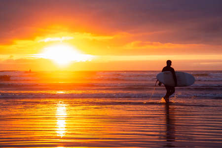 Surfer walking towards the Ocean on the sandy beach during a dramatic summer sunset. Taken in Seaside, Oregon, United States of America.の写真素材