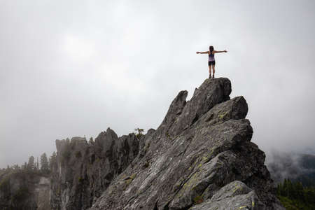 Adventurous Girl with Open Arms on top of a rugged rocky mountain during a cloudy summer morning. Taken on Crown Mountain, North Vancouver, BC, Canada.の写真素材