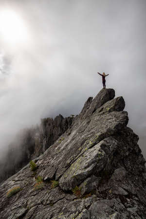 Adventurous Girl with Open Arms on top of a rugged rocky mountain during a cloudy summer morning. Taken on Crown Mountain, North Vancouver, BC, Canada.の写真素材