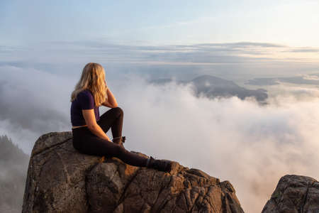Adventurous Female Hiker on top of a mountain covered in clouds during a vibrant summer sunset....