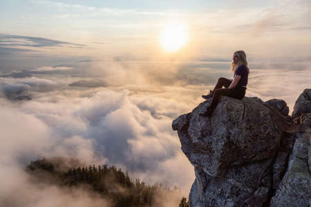 Adventurous Female Hiker on top of a mountain covered in clouds during a vibrant summer sunset. Taken on top of St Mark's Summit, West Vancouver, British Columbia, Canada.の写真素材
