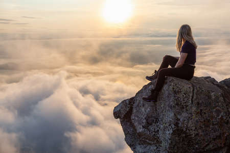 Adventurous Female Hiker on top of a mountain covered in clouds during a vibrant summer sunset. Taken on top of St Marks Summit, West Vancouver, British Columbia, Canada.の写真素材