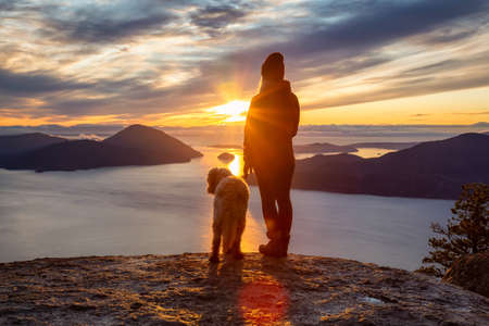 Adventurous Girl Hiking on top of a Mountain with a dog during a colorful sunset. Taken on Tunnel Bluffs Hike, near Vancouver and Squamish, British Columbia, Canada.の写真素材