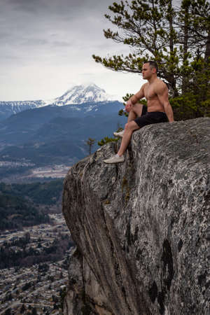 Fit and Muscular Caucasian Man is sitting at the cliff on top of a mountain and enjoying the scenery. Taken in Squamish, North of Vancouver, BC, Canada.の写真素材