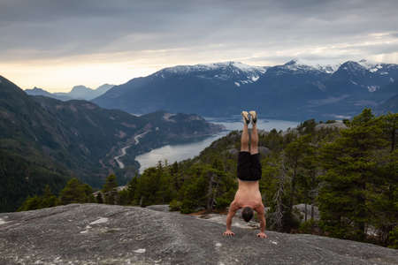 Fit and Muscular Young Man is doing exercises on top of the Mountain during a cloudy sunset. Taken...